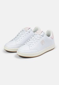 White leather sneakers with a smooth texture, featuring pink accents on the heel and tongue, round toe, and white laces. Brown rubber outsole.