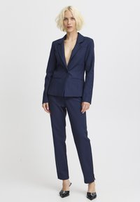 Námorně modrý oblek na míru s blazrem s hlubokým V-výstřihem a dvěma předními kapsami, doplněný o slim-fit kalhoty a špičaté černé boty.