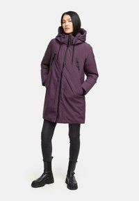 Lilla isoleret parka med hætte, der har en fuld frontlynlås, sidelommer og justerbare snore. Båret over sorte tætsiddende bukser og støvler.