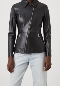 Černá kožená bunda s límcem, asymetrickým zipem a dvěma zipovými postranními kapsami. Hladká textura a přiléhavý design v kombinaci se světlými džíny.