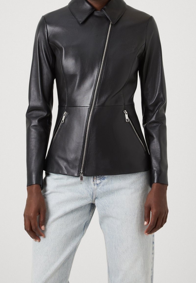 Černá kožená bunda s límcem, asymetrickým zipem a dvěma zipovými postranními kapsami. Hladká textura a přiléhavý design v kombinaci se světlými džíny.