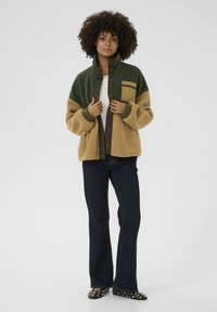 Giacca in pile verde e beige con tasca frontale; presenta un colletto alto e bottoni a pressione. Abbinata a jeans svasati scuri e scarpe con fantasia.