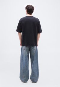 Άνδρας που στέκεται με την πλάτη προς τα εμπρός, φορώντας μπλούζα μαύρου χρώματος και φαρδιά ξεθωριασμένα μπλε τζιν, μπροστά σε απλό λευκό φόντο.