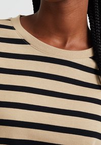 Detailní záběr osoby oble čené v béžovém a černém pruhovaném tričku s copánky viditelnými na pravé straně.