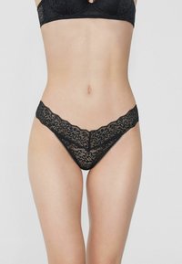 Schwarzer Spitzen-Tanga mit aufwändigen Blumenmustern, gewellten Kanten und einer leichten, halbtransparenten Textur für ein zartes Erscheinungsbild.