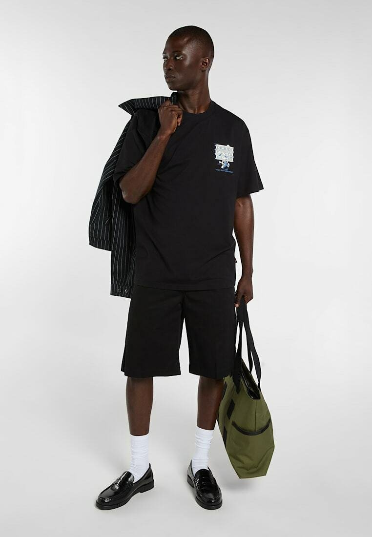 Junger Mann trägt schwarzes T-Shirt und Shorts, hält eine grüne Tragetasche und einen Nadelstreifen-Blazer über der Schulter, steht vor einfachem Hintergrund.