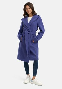 Cappotto viola con cappuccio e cintura, maniche lunghe, tasche laterali e una texture morbida. Indossato sopra un top bianco e abbinato a jeans e sneakers.