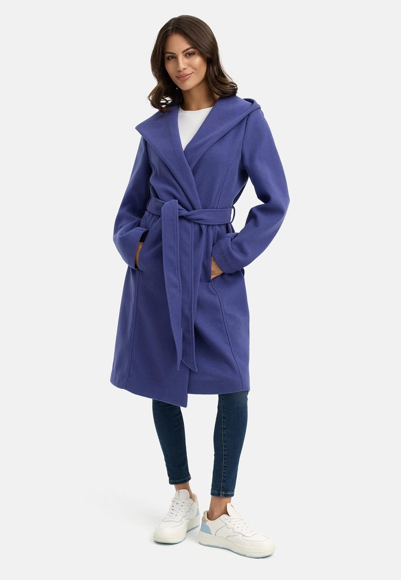 Cappotto viola con cappuccio e cintura, maniche lunghe, tasche laterali e una texture morbida. Indossato sopra un top bianco e abbinato a jeans e sneakers.