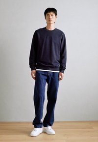 Tmavomodrá mikina z bavlny, skombinovaná s voľnými modrými džínsami. Biele tenisky dotvárajú outfit, na jednoliatom pozadí.