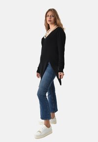 Top nero a maniche lunghe con scollatura a V e orlo appuntito, abbinato a jeans a zampa d'elefante blu e sneaker bianche con dettagli dorati.
