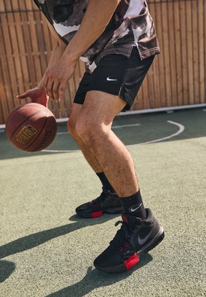 Osoba dryblująca piłkę do koszykówki na zewnętrznym boisku, ubrana w czarne spodenki Nike, czarno-czerwone buty do koszykówki i czarne skarpetki Nike.