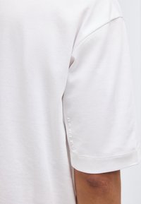 Κοντινό πλάνο λευκού κοντομάνικου πουκάμισου σε άτομο, που δείχνει το πάνω μέρος του βραχίονα και ένα μικρό κάθετο λογότυπο "Oakley" κοντά στην άκρη του μανικιού.