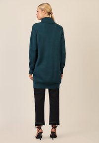 Teal striktrøje med høj krave, lange ærmer og ribbede kanter. Kombinationen med sorte cropped jeans med frynset kant og stroppehæle.