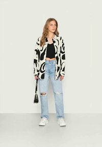 Camisa oversized em preto e branco com padrão, top cropped preto, jeans rasgados em azul claro, ténis brancos e uma mala preta.