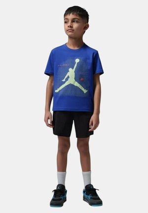 Junge steht und trägt ein blaues T-Shirt mit einer Basketball-Silhouette, schwarze Shorts, weiße Socken und schwarze blaue Sneaker.