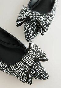 Juodi aštrūs balerinos bateliai, dekoruoti daugybe mažų sidabro spalvos studų ir dideliu kaspinu su atitinkamais akcentais. Lygus paviršius.