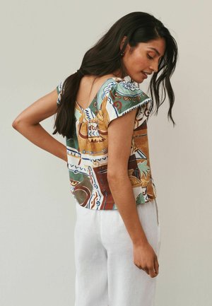 Kobieta z długimi ciemnymi włosami ubrana w bluzkę z krótkim rękawem w wzory oraz białe spodnie, patrząca w dół z ręką na biodrze na jednolitym tle.