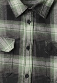 Chemise à carreaux verte, noire et blanche à boutons avec un col et deux poches avant, dont une avec un écusson noir en forme de losange cousu.