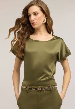 Vrouw met lang golvend haar, gekleed in een olijfgroene satijnen top met korte mouwen, bijpassende broek, gouden oorbellen en een riem met patroon, met handen in de zakken.