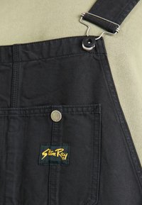 Zwarte katoenen salopettes hebben een voorvak met een zilveren drukknoop en een Stan Ray geborduurd logo in het geel op een groene achtergrond.
