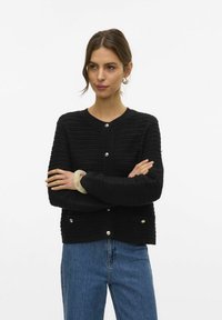 Donna con capelli castani che indossa un cardigan nero a bottoni con texture, jeans blu, orecchini a cerchio argentati e un grande braccialetto beige, con le braccia incrociate.