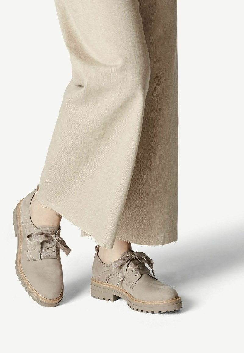 Chaussures beiges en daim à lacets avec semelle épaisse portées avec un pantalon large beige clair coupé au-dessus de la cheville, sur fond blanc.