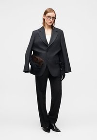 Blazer oversize gris anthracite à fines rayures avec larges épaules, pantalon noir et chaussures noires élégantes. Tenant une pochette marron.