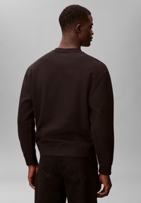 Fekete hosszú ujjú pulóvert és fekete nadrágot viselő férfi, hátat fordítva, egyszerű szürke háttér előtt áll.