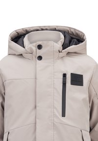 Beige Winterjacke mit Kapuze, ausgestattet mit einer Reißverschlusstasche auf der Brust, Druckknöpfen und einem strukturierten Stoff mit matter Oberfläche.