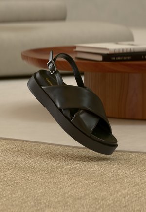 Sandalia negra de cuero con diseño de tiras cruzadas y hebilla. Suela negra gruesa con ligera textura, suspendida sobre una superficie beige texturizada.