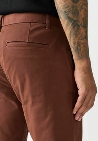 Braune Baumwollhosen mit glatter Textur, ausgestattet mit einer Gesäßtasche und tailliertem Schnitt. Die Seitenansicht zeigt eine schlanke Silhouette und Gürtelschlaufen.