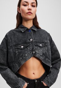 Veste en denim noir coupée avec un col, deux poches frontales et une fermeture à bouton. Caractéristiques d'une texture délavée et d'un bouton d'accent bleu.