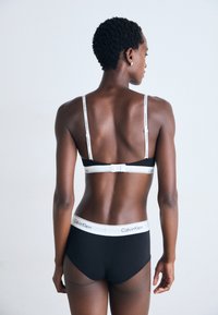Svart Calvin Klein-bh og høyhælte truser, med hvite logobånd og livlinning. Elegant, minimalistisk design med glatt tekstur.