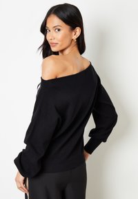 Zwart off-the-shoulder trui met lange, ballonmouwen, geribbelde zoom en een textuur brei. Glad materiaal tegen een lichte achtergrond.