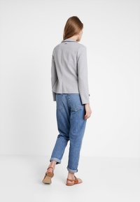 Blazer gris ajustado con una estructura entallada, exhibido sobre unos pantalones vaqueros denim azules holgados, complementado con sandalias planas en tono marrón.