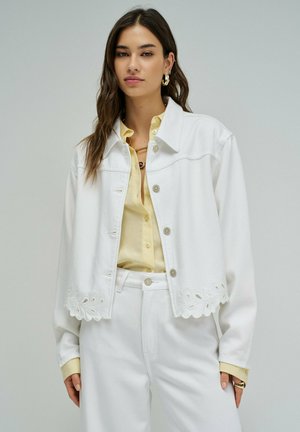 Mujer con chaqueta blanca abotonada con borde festoneado sobre una camisa amarillo pálido y pantalones blancos, contra un fondo gris liso.