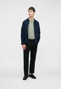 Camicia di lino blu navy, maglione a collo rotondo verde chiaro, pantaloni neri e scarpe nere. Il modello si trova su uno sfondo uniforme, guardando verso l'alto.