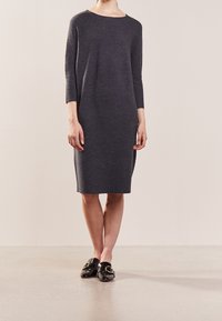 Donna in piedi, che indossa un vestito grigio scuro al ginocchio, con maniche lunghe e scarpe slip-on in pelle sintetica nera con cinturini dorati.