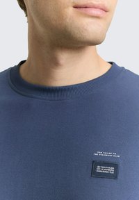 Niebieska bluza z prążkowanym kołnierzem, z prostokątną naszywką z logo i tekstem. Tkanina jest gładka i miękka w dotyku.