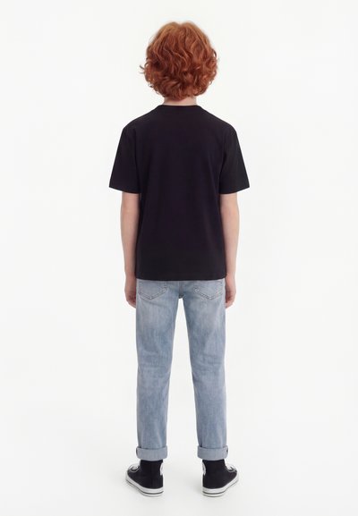 Παιδί με σγουρά κόκκινα μαλλιά, γυρισμένο προς τα πίσω, φοράει μαύρο μπλουζάκι, ανοιχτό μπλε τζιν με γυρισμένα μπατζάκια και μαύρα ψηλά αθλητικά παπούτσια.