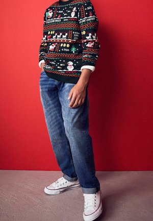 Personne portant un pull festif avec des motifs sur le thème des jeux, un jean bleu et des baskets blanches, debout devant un mur rouge.