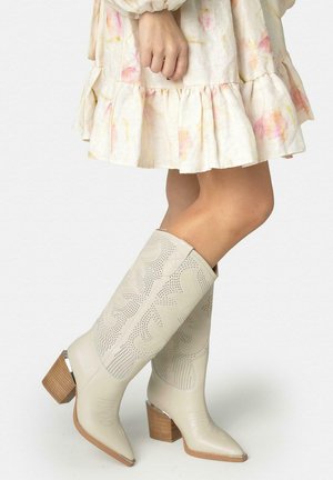 Mujer con botas de cuero beige hasta la rodilla, con tacones de madera y bordados, combinadas con un vestido claro con flores y volantes.