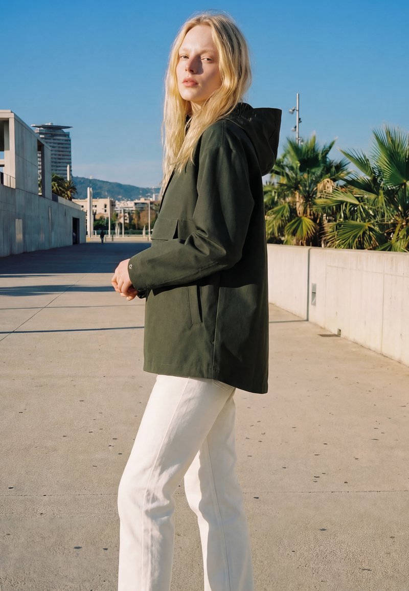 Giovane donna con lunghi capelli biondi che indossa una giacca con cappuccio verde scuro e pantaloni bianchi, in piedi all'aperto in una giornata di sole.