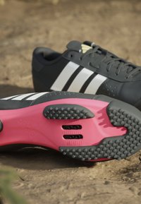 Černá sportovní bota s bílými pruhy. Texturovaná gumová podrážka v růžové barvě, s malými hroty a odvodňovacími otvory. Hladký svrchní materiál.