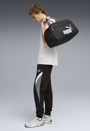 Ung mand i hvid T-shirt og sorte Puma træningsbukser med en sort Puma duffel taske over skulderen, stående foran en grå baggrund.