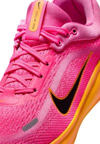 Rožu sporta apavi ar teksturētu sieta augšdaļu, melnu Nike zīmi, dzeltenu zoli un rožainām šņorēm; iekļauts uzraksts "STELLAR RIDE".