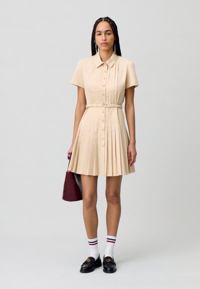 Vestido beige de mangas cortas con cuello, con botones frontales y pliegues. Combinado con mocasines negros y calcetines blancos con acentos rojos y azules.