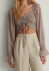 Crop top i ljus taupe med rynkad framsida, dragsko-detalj och genomskinliga långa ärmar. Matchas med högmidjade beige byxor.