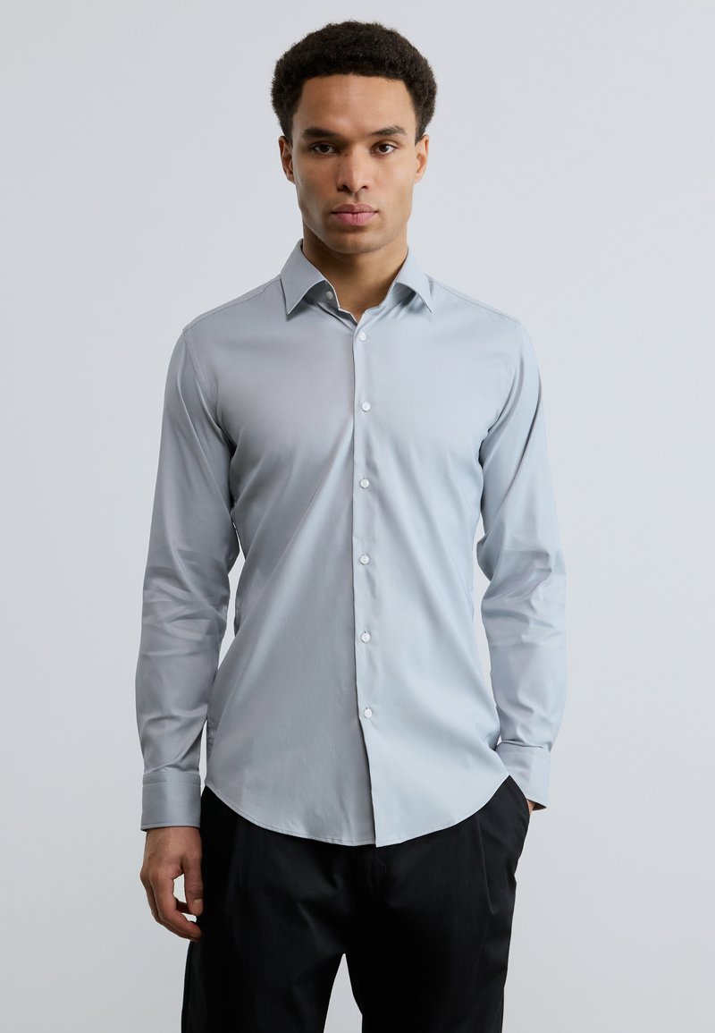 Άνδρας που στέκεται με το πρόσωπο προς τα εμπρός φορόντας ανοιχτό γκρι πουκάμισο με κουμπιά μακρυμάνικο και μαύρο παντελόνι, με το ένα χέρι στην τσέπη.