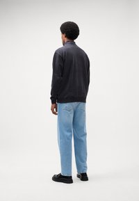 Sort, lynlåset sweatshirt med en blød tekstur, parret med lyseblå, afslappede jeans og sorte sko. Personen ses fra ryggen.